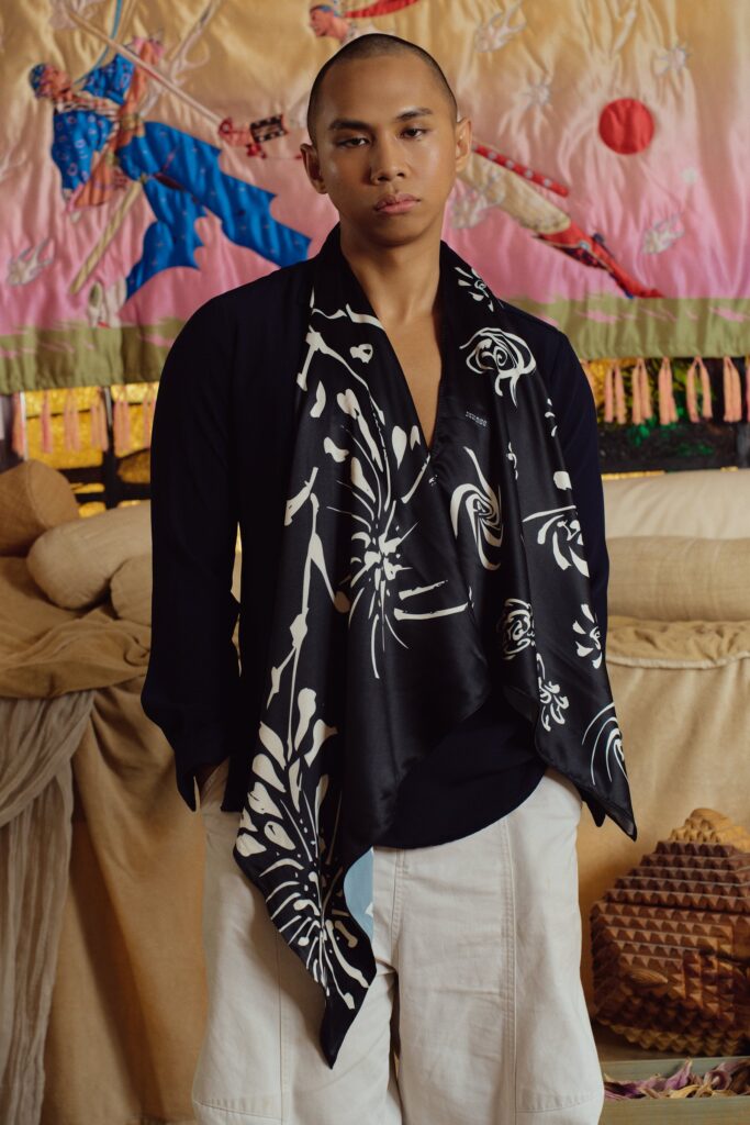 Model wearing the black Flower diamond scarf draped over a dark jacket, quilted tapestry behind